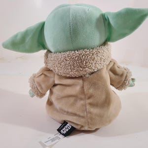 May include: A plush toy of a character from the Star Wars franchise, featuring a light green head and large ears. The toy is wearing a tan coat with a fuzzy collar and cuffs. The toy is sitting down.