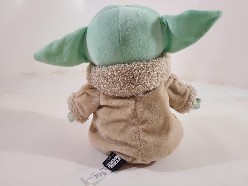 May include: A plush toy of a character from the Star Wars franchise, featuring a light green head and large ears. The toy is wearing a tan coat with a fuzzy collar and cuffs. The toy is sitting down.