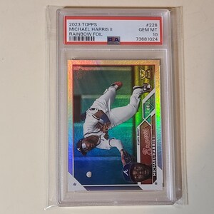 May include: A baseball card featuring Michael Harris II of the Atlanta Braves in a rainbow foil design. The card is graded GEM MT 10 and encased in a protective holder. The card's text includes "2023 Topps" and "Rainbow Foil".