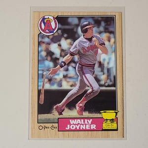 May include: A vintage O-Pee-Chee baseball card featuring Wally Joyner in a grey Angels uniform, swinging a bat. The card has a wood-grain border, the Angels logo in the top left, and a yellow trophy icon with the text All Star Rookie in the corner.