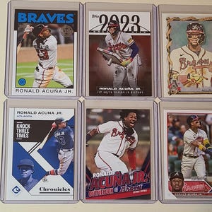 May include: Six baseball trading cards featuring Ronald Acuña Jr. in various poses and uniforms. The cards are encased in protective sleeves. The top row includes cards with the Braves logo and the text "2023".