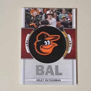 May include: Baseball trading card featuring Adley Rutschman of the Baltimore Orioles. The card has a black and orange Orioles logo, the team's initials "BAL", and the player's name. The background shows a baseball game.