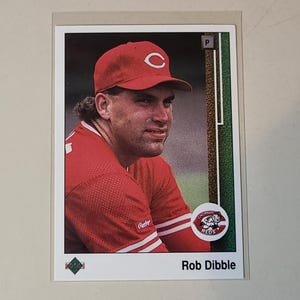 May include: Baseball card featuring Rob Dibble in a red Cincinnati Reds uniform and cap. The card has the team logo and Dibble's name printed at the bottom. The background is a white border.