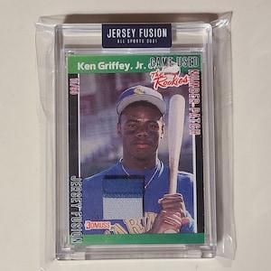 May include: A baseball card featuring Ken Griffey Jr. in a blue jersey with a white and blue jersey patch. The card is from the Jersey Fusion All Sports 2021 collection and features the text "Game Used Number Patch" and "The Rookies".