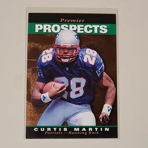 Puede incluir: Una tarjeta de intercambio de fútbol americano con Curtis Martin, un corredor de los New England Patriots. La tarjeta es de la serie Premier Prospects y presenta una foto de Martin con su uniforme sosteniendo un balón de fútbol. La tarjeta tiene un fondo verde y dorado con el texto "Premier Prospects" y "Curtis Martin" en blanco.
