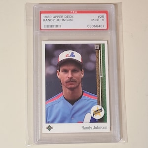 May include: A 1989 Upper Deck Randy Johnson baseball card, graded PSA Mint 9, encased in a clear plastic protective slab. The card features a portrait of the player wearing a light blue jersey and a white cap with a red and blue logo.
