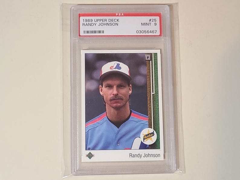 May include: A 1989 Upper Deck Randy Johnson baseball card, graded PSA Mint 9, encased in a clear plastic protective slab. The card features a portrait of the player wearing a light blue jersey and a white cap with a red and blue logo.