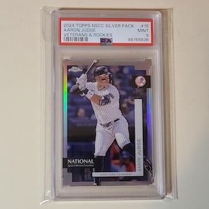May include: A baseball card of Aaron Judge in a protective case. The card is from the 2024 Topps NSCC Silver Pack, featuring Judge in a New York Yankees uniform. The card is graded as Mint 9 by PSA.