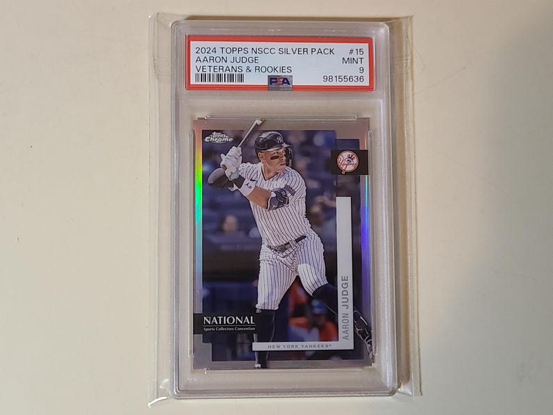 May include: A baseball card of Aaron Judge in a protective case. The card is from the 2024 Topps NSCC Silver Pack, featuring Judge in a New York Yankees uniform. The card is graded as Mint 9 by PSA.