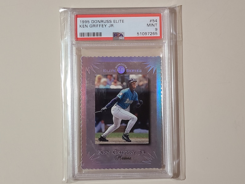 May include: A 1995 Donruss Elite Series baseball card featuring Ken Griffey Jr. of the Mariners. The card is graded 9 by PSA and has a holographic finish.