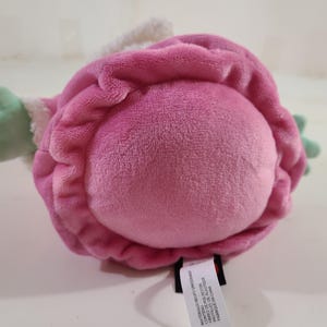 Pu&ograve; includere: Un giocattolo di peluche a forma di fiore, principalmente rosa con accenti verdi. Il giocattolo ha una consistenza morbida e vellutata e un bordo bianco. Il giocattolo &egrave; appoggiato su una superficie bianca.