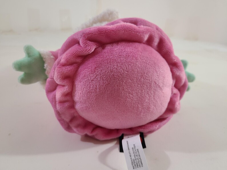 Pu&ograve; includere: Un giocattolo di peluche a forma di fiore, principalmente rosa con accenti verdi. Il giocattolo ha una consistenza morbida e vellutata e un bordo bianco. Il giocattolo &egrave; appoggiato su una superficie bianca.