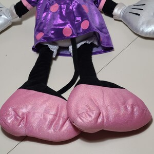 Pu&ograve; includere: Un giocattolo di peluche a forma di topo dei cartoni animati che indossa un vestito viola con pois rosa e scarpe rosa. Il giocattolo ha gambe nere e guanti argentati.