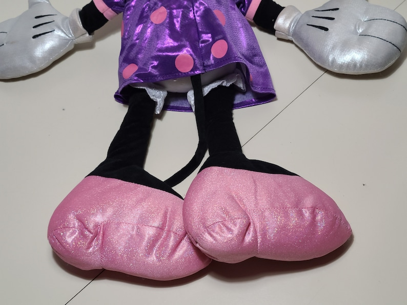 Pu&ograve; includere: Un giocattolo di peluche a forma di topo dei cartoni animati che indossa un vestito viola con pois rosa e scarpe rosa. Il giocattolo ha gambe nere e guanti argentati.