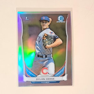 May include: A baseball card featuring a baseball player in a blue and white uniform with a red "C" on the chest. The card is a Bowman Chrome 1st Edition card and features Dylan Cease, a pitcher. The card has a holographic background.
