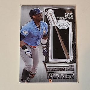 May include: A baseball card featuring Yandy Diaz, First Baseman, in a Tampa Bay Rays uniform. The card includes a Louisville Slugger logo and text indicating a 2023 Silver Slugger Award Winner. The background is a gradient of silver and gray.