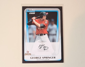 May include: A baseball card of George Springer, Houston Astros. The card has a blue and white border with "1st Bowman Card" at the top. Springer is in a batting stance, wearing a red jersey and white trousers. His signature is visible.