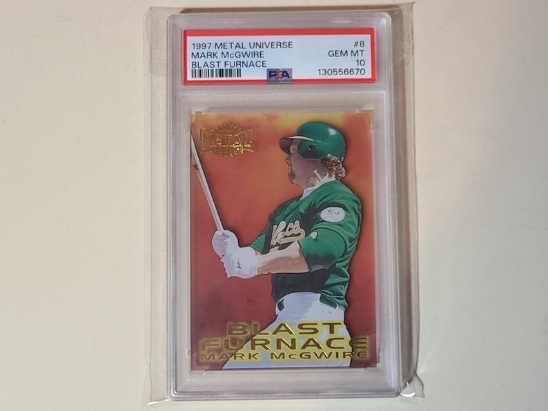 May include: A baseball card featuring Mark McGwire in a green and white uniform, holding a bat. The card is from the 1997 Metal Universe series, titled "Blast Furnace." The card is encased in a protective holder with text that reads "GEM MT 10."