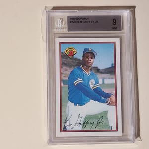 Peut inclure: Une carte de baseball Bowman de 1989 de Ken Griffey Jr. est enferm&eacute;e dans un support de protection. La carte pr&eacute;sente une image couleur de Griffey en uniforme de baseball bleu. La note Beckett indique un &eacute;tat "9" parfait.