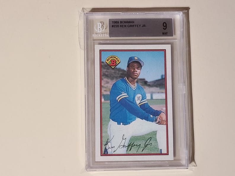 Peut inclure: Une carte de baseball Bowman de 1989 de Ken Griffey Jr. est enferm&eacute;e dans un support de protection. La carte pr&eacute;sente une image couleur de Griffey en uniforme de baseball bleu. La note Beckett indique un &eacute;tat "9" parfait.