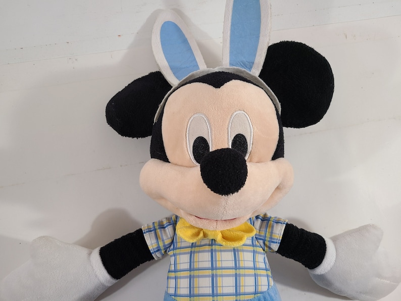 May include: A plush Mickey Mouse toy wearing blue bunny ears and a yellow and blue plaid shirt with a yellow bow tie.