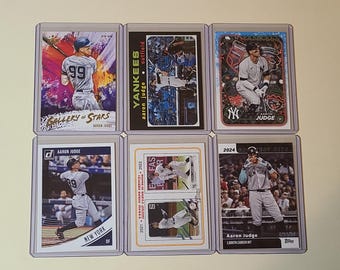 May include: Six baseball trading cards featuring Aaron Judge of the New York Yankees. The cards are in protective sleeves and showcase various action shots and designs. The cards include text such as "Gallery of Stars" and "2024 Greatest Hits."