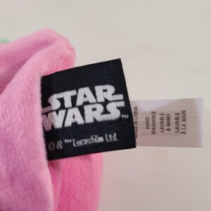 Pu&ograve; includere: Un articolo in peluche rosa con un'etichetta nera di Star Wars e un'etichetta bianca con istruzioni di lavaggio. Il logo di Star Wars &egrave; bianco. L'etichetta include le parole "Lavabile a mano" e "Made in China."