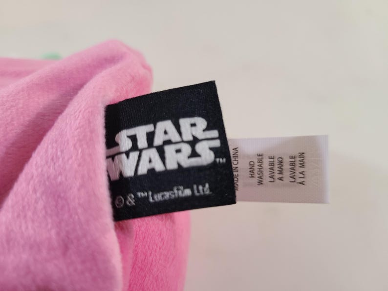 Pu&ograve; includere: Un articolo in peluche rosa con un'etichetta nera di Star Wars e un'etichetta bianca con istruzioni di lavaggio. Il logo di Star Wars &egrave; bianco. L'etichetta include le parole "Lavabile a mano" e "Made in China."