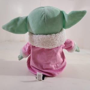 Pu&ograve; includere: Un peluche di un personaggio con grandi orecchie e un abito rosa. Il giocattolo ha una testa e orecchie verde menta, un indumento rosa simile a una veste e un colletto bianco e soffice. Il giocattolo &egrave; progettato per assomigliare a un personaggio popolare.
