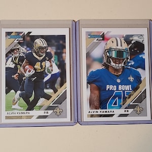 May include: Two 2019 Donruss football trading cards featuring Alvin Kamara. The left card shows Kamara in a black uniform running with a football. The right card shows Kamara in a blue Pro Bowl uniform with a helmet visor. Both cards are in plastic cases.