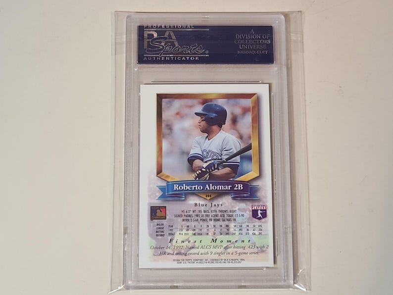 May include: A professionally graded Roberto Alomar baseball card encased in a clear plastic PSA holder. The card features a photograph of the player in a Blue Jays uniform, with text detailing his stats, position as 2B, and his 1992 ALCS MVP award.