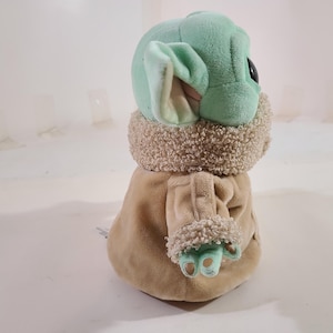May include: A plush toy of a character with large ears, green skin, and a tan coat. The toy has a light green head and ears, with a tan coat and a fluffy, light brown collar. The character's hands and feet are light green.