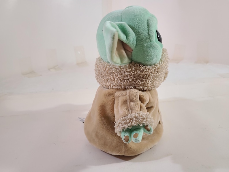 May include: A plush toy of a character with large ears, green skin, and a tan coat. The toy has a light green head and ears, with a tan coat and a fluffy, light brown collar. The character's hands and feet are light green.
