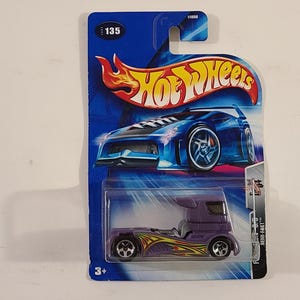May include: A purple Hot Wheels Semi-Fast 1:64 scale toy truck with yellow flames on the side. The truck is on a blue and silver card with the Hot Wheels logo and the number 135.