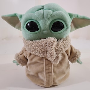 May include: A plush doll of a character with large black eyes, large ears, and a light green complexion. The doll wears a tan coat with a fluffy collar and cuffs. The doll is a popular character from a science fiction series.