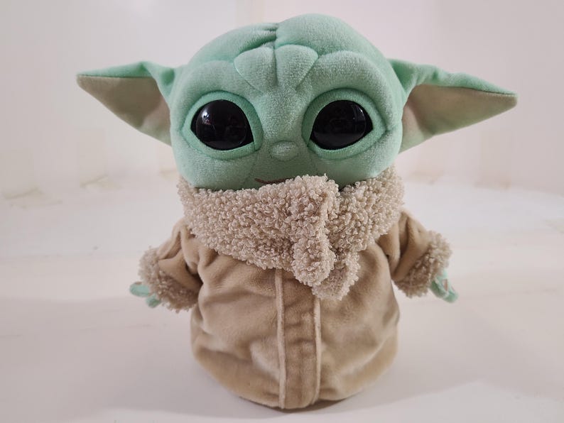 May include: A plush doll of a character with large black eyes, large ears, and a light green complexion. The doll wears a tan coat with a fluffy collar and cuffs. The doll is a popular character from a science fiction series.