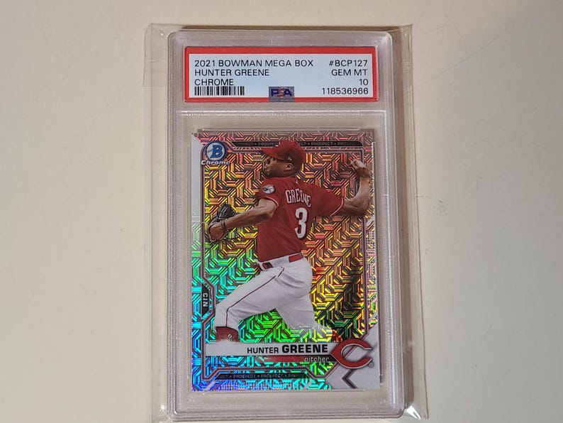 May include: A 2021 Bowman Mega Box Hunter Greene Chrome baseball card, graded PSA Gem Mt 10, #BCP127. The card features a holographic, geometric pattern background with the pitcher in a red jersey and white pants, encased in a clear plastic slab.