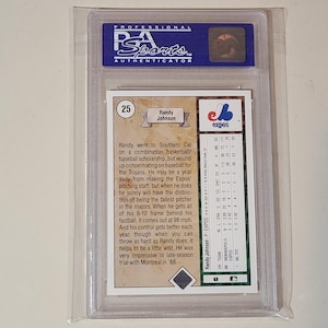 May include: A PSA graded baseball card featuring Randy Johnson, encased in a clear plastic holder. The back of the card displays biographical text, the number 25, and a statistical table for the Montreal Expos, including team and pitching data.