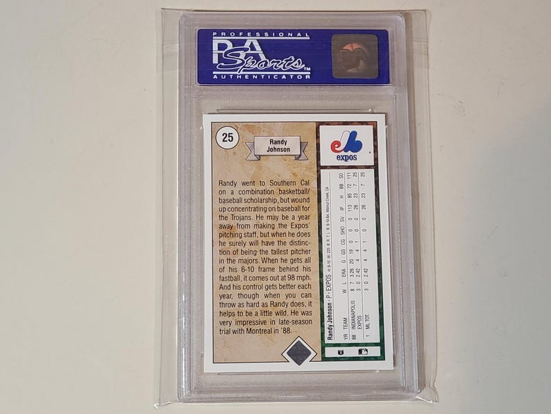 May include: A PSA graded baseball card featuring Randy Johnson, encased in a clear plastic holder. The back of the card displays biographical text, the number 25, and a statistical table for the Montreal Expos, including team and pitching data.