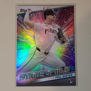 May include: A baseball card featuring Paul Skenes of the Pittsburgh Pirates in a pitching pose. The card has a holographic design with the text "STARS OF MLB" and "PAUL SKENES" at the bottom. The Topps logo is at the top.