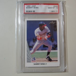 Puede incluir: Una tarjeta de b&eacute;isbol clasificada con Sammy Sosa, un ex jugador de los Chicago White Sox. La tarjeta es de la serie Leaf de 1990 y est&aacute; clasificada GEM MT 10 por PSA. La tarjeta muestra a Sosa en posici&oacute;n de bateo, vistiendo un uniforme blanco y rojo con el logotipo de los Chicago White Sox. La tarjeta est&aacute; en una caja protectora.