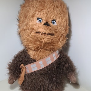 May include: A brown and tan plush toy of Chewbacca from Star Wars. The toy has blue eyes and a brown fur vest with a white bandolier.