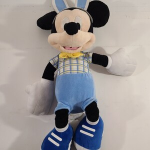 May include: A plush Mickey Mouse toy wearing blue bunny ears, a yellow bow tie, and a blue and yellow plaid shirt. He is wearing blue pants and blue sneakers with white accents.