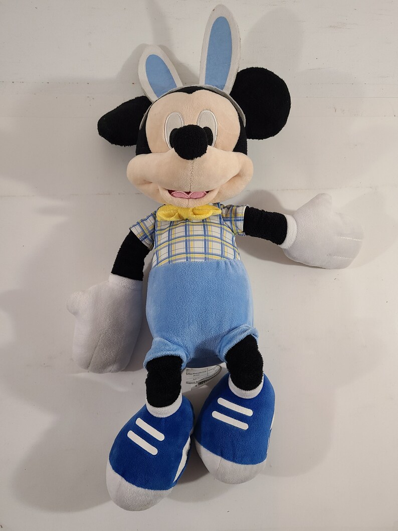 May include: A plush Mickey Mouse toy wearing blue bunny ears, a yellow bow tie, and a blue and yellow plaid shirt. He is wearing blue pants and blue sneakers with white accents.