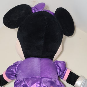 Pu&ograve; includere: Un giocattolo di peluche di Minnie Mouse con un vestito di raso viola con guanti di paillettes argentati. Le sue orecchie sono nere e il suo viso &egrave; bianco.