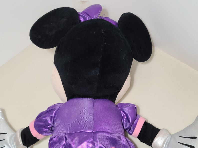 Pu&ograve; includere: Un giocattolo di peluche di Minnie Mouse con un vestito di raso viola con guanti di paillettes argentati. Le sue orecchie sono nere e il suo viso &egrave; bianco.