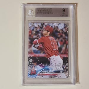 May include: A baseball card featuring Shohei Ohtani in a red Angels uniform, with a snowflake design. The card is graded and encased in a clear plastic holder. The card is labeled "2018 Topps Walmart Holiday Snowflake".