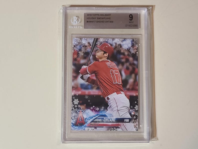 May include: A baseball card featuring Shohei Ohtani in a red Angels uniform, with a snowflake design. The card is graded and encased in a clear plastic holder. The card is labeled "2018 Topps Walmart Holiday Snowflake".