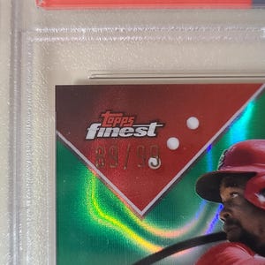 K&ouml;nnte beinhalten: Baseball-Sammelkarte mit einem Spieler in rotem Helm und Uniform. Die Karte hat einen roten und gr&uuml;nen Hintergrund mit dem Text "Topps Finest" und der Nummer "89/99". Die Karte ist in einem Schutzhalter eingeschlossen.