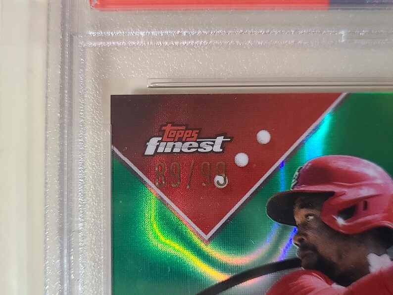 K&ouml;nnte beinhalten: Baseball-Sammelkarte mit einem Spieler in rotem Helm und Uniform. Die Karte hat einen roten und gr&uuml;nen Hintergrund mit dem Text "Topps Finest" und der Nummer "89/99". Die Karte ist in einem Schutzhalter eingeschlossen.